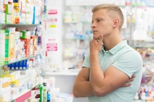 Man standing in pharmacy in front of shelf full of different medicines trying to pick one