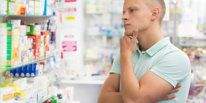 Man standing in pharmacy in front of shelf full of different medicines trying to pick one